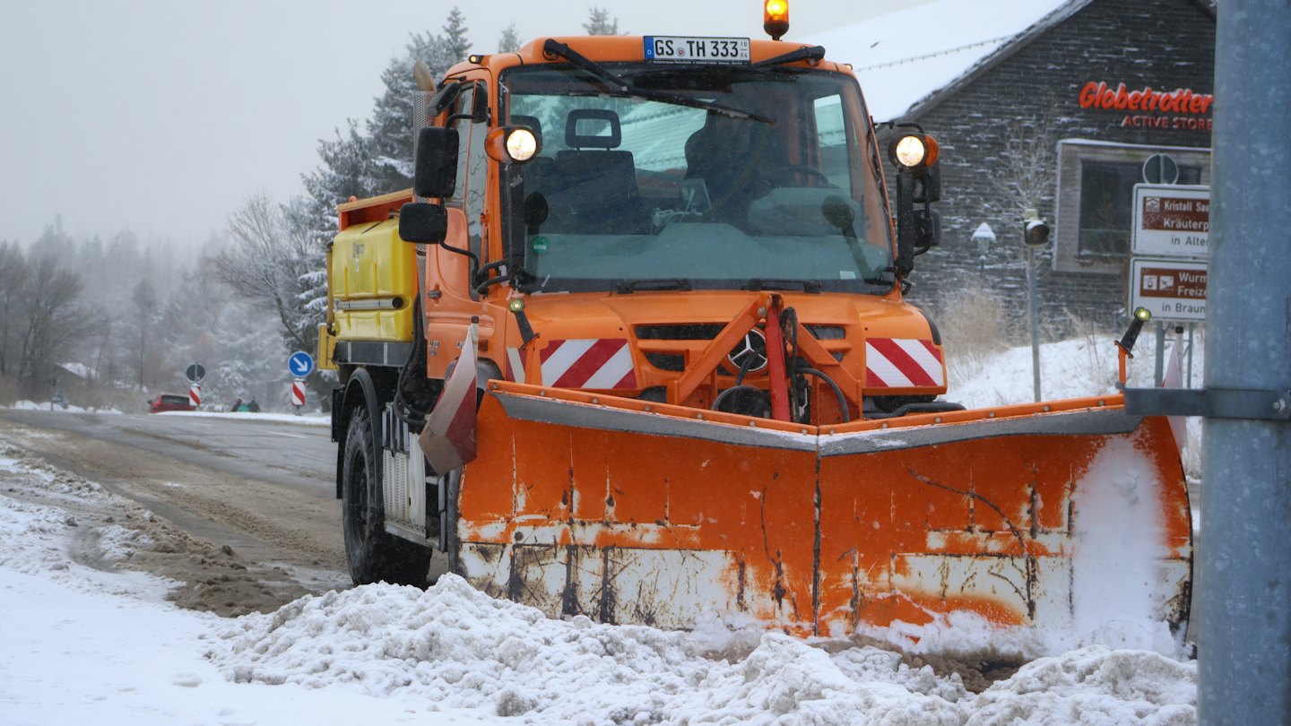 Orangefarbener Schneepflug räumt eine verschneite Straße.